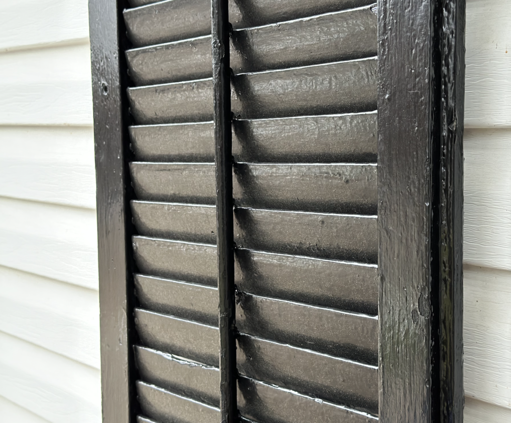 A closeup of a painted black shutter shows old paint, slats, and a push rod. 